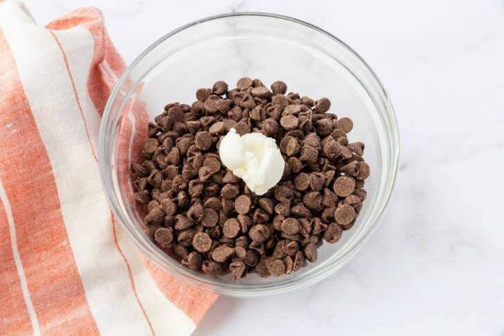 A glass bowl filled with chocolate chips and a dollop of white butter sits on a marble surface. An orange and white striped cloth is partially visible on the left side of the image.