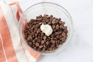 A glass bowl filled with chocolate chips and a dollop of white butter in the center. The bowl is placed on a white surface next to an orange and white striped cloth.