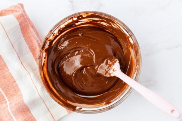 A bowl of melted chocolate with a pink spatula, placed next to an orange and white striped kitchen towel on a white marble surface.