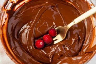 A fork holding three red berries rests in a bowl of melted chocolate, creating a rich and delectable scene. The smooth surface of the chocolate contrasts with the vibrant color of the berries.