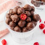 A glass bowl filled with chocolate-covered cherries. One cherry is cut in half to show the red center. More cherries and a striped cloth are in the background on a white surface.