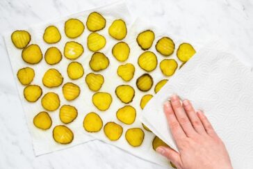 Hand placing a paper towel over rows of sliced pickles on a white marble surface.