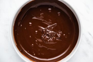A white bowl filled with smooth, glossy dark chocolate ganache on a light marble surface.