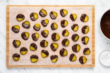A wooden board holds several slices of chocolate-dipped dried pineapple, arranged in rows. The pineapples are partially covered in dark chocolate, and there's a bowl of chocolate and small bowl of salt to the side.