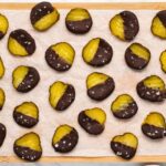 Pickle slices partially dipped in chocolate are arranged on a parchment-lined tray. Some are sprinkled with salt flakes.