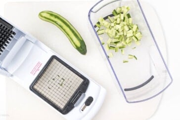 A vegetable dicer with a grid blade is shown in use. Diced cucumber pieces are in a clear container, and a whole cucumber is next to the dicer on a light-colored cutting board.