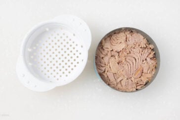 An opened can of tuna is displayed next to a white plastic strainer on a speckled white surface. The tuna is light in color and chunky, filling the entire can.