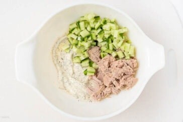 A white bowl containing diced cucumber, flaked tuna, and a creamy mayonnaise-based dressing with herbs, placed on a light surface.