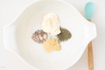 A white bowl with mayonnaise, ground black pepper, dried dill, garlic powder, and mustard. A wooden spoon with a turquoise handle rests on the side of the bowl. The background is a white, speckled surface.