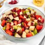 A bowl of colorful fruit salad with diced apples, pears, strawberries, kiwi, grapes, and pomegranate seeds, set on a white surface. There's a small bowl of pomegranate seeds and a halved lemon in the background.