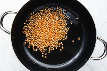 A black pan on a marble surface filled with unpopped popcorn kernels and a small amount of oil. The kernels are evenly spread across the bottom, ready for popping.