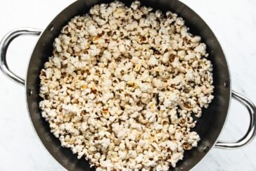 A large pot filled with freshly popped popcorn. The popcorn is evenly distributed and appears fluffy and lightly browned, sitting against a white marble background. The pot has two handles on either side.