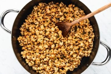 A large pot filled with freshly popped popcorn coated in a golden seasoning, with a wooden spoon resting inside. The pot has metal handles on both sides and is placed on a light surface.