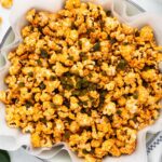 A bowl of spicy popcorn garnished with chopped jalapeños on a parchment-lined dish. The popcorn is seasoned with a reddish-brown spice mix, giving it a bold, flavorful appearance. The edges of the parchment paper peek out from the bowl.