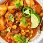 A close-up of a bowl of stew featuring chunks of potato, corn, black beans, tomatoes, and green peppers. Garnished with fresh cilantro, sliced jalapeño, and a lime wedge. A spoon is placed in the bowl.