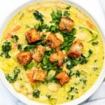 A bowl of creamy soup contains white beans, kale, carrots, and celery, garnished with crispy croutons and chopped parsley. The soup has a light yellow base, suggesting a rich, flavorful broth.