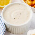 A white ramekin filled with creamy, seasoned white sauce is placed next to a lemon wedge, a striped cloth, and a dish of golden fries.