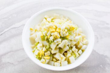 A white bowl filled with chopped hard-boiled eggs and celery pieces, placed on a light marbled surface. The ingredients are mixed together, showcasing a simple, homemade salad or dish preparation.
