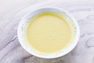 A white bowl filled with creamy yellow soup, placed on a light marbled surface. The soup appears smooth, garnished with specks of herbs, and the bowl's rim is clean, emphasizing the dish's rich texture.