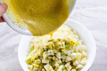 A bowl of chopped hard-boiled eggs and diced celery is being covered with creamy dressing poured from a bowl.