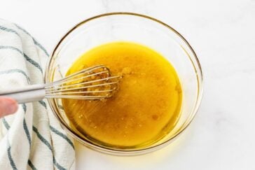 A glass bowl filled with golden liquid, likely a dressing or marinade, is being whisked by a hand holding a metal whisk. A white and green striped cloth is partially visible on the side.