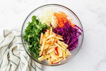 A glass bowl filled with shredded green kale, white cabbage, orange carrots, purple cabbage, and sliced apples. A striped cloth is placed beside the bowl on a marble surface.
