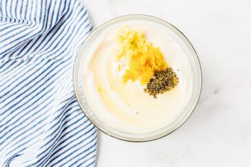 A clear bowl of creamy white yogurt on a marble surface, topped with grated lemon zest, minced garlic, and cracked black pepper. A blue and white striped kitchen towel is partially visible on the left.