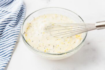 A clear glass bowl filled with creamy, speckled dressing. A metal whisk rests inside the bowl. A blue and white striped cloth is partially visible on the left side on a white marble surface.