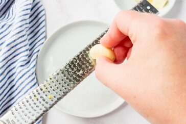 A hand grates a clove of garlic on a metal grater over a white plate. A blue and white striped cloth is visible to the side.