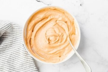 A bowl of creamy orange dip with a smooth texture, set on a marble surface next to a striped cloth. A spoon rests in the bowl, indicating it's ready to be served or used.