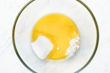 A clear glass mixing bowl on a marble surface contains melted butter, a scoop of sugar, and a small amount of baking powder, ready for mixing.