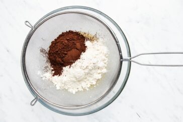 A sifter containing cocoa powder and flour sits on a marble countertop, ready for baking preparation.