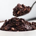 A close-up of a spoon scooping rich chocolate cookie dough with chunks of chocolate from a white bowl. The dough looks creamy and dense, with a glossy texture. The background is softly blurred.
