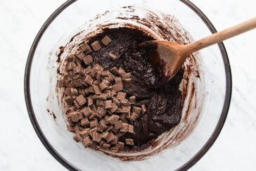 A glass bowl containing chocolate chip brownie batter, with a wooden spoon resting inside. The batter is thick and dark, and the chocolate chips are scattered on top, ready to be mixed in. The bowl is set on a marble surface.