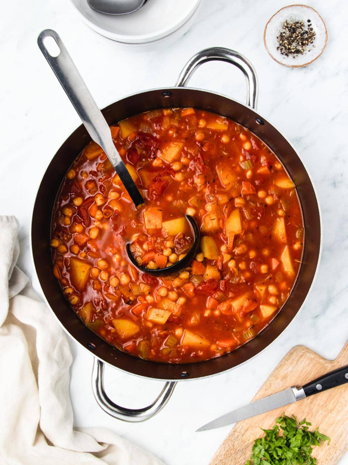 A pot of hearty vegetable stew with chickpeas, potatoes, and tomatoes, accompanied by a ladle. The stew is placed on a marble surface next to chopped herbs on a wooden board and a small dish of pepper.