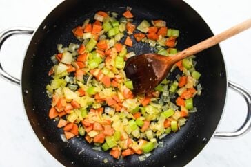 A black skillet contains diced carrots, celery, and onions being sautéed. A wooden spoon rests inside the skillet, stirring the colorful vegetables. The background is a white countertop.
