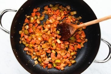 A black pan with chopped carrots, green bell peppers, and onions being sautéed. A wooden spoon rests in the pan, stirring the colorful vegetables.
