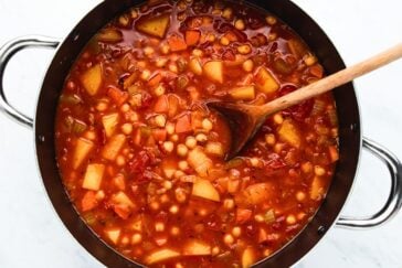 A large pot filled with a colorful vegetable soup. The soup contains chunks of potatoes, carrots, and chickpeas, along with diced tomatoes and a rich broth. A wooden spoon is stirring the mixture.