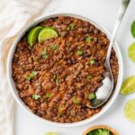 A bowl of hearty chili made with black beans and garnished with fresh cilantro and lime wedges. A spoon rests inside the bowl. A light cloth and lime slices surround the dish on a white surface.