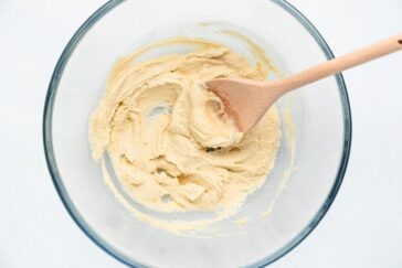 A glass bowl containing a creamy, light beige batter is being mixed with a wooden spoon. The batter has a smooth consistency and is well blended. The background is a plain white surface.