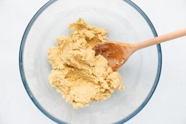 A glass bowl containing light brown cookie dough with a wooden spoon resting in the mixture, set on a light background.