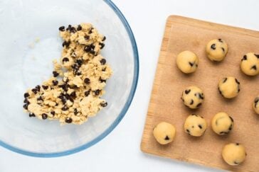 A glass bowl with chocolate chip cookie dough sits on the left, partially mixed. On the right, a wooden tray holds several rounded balls of dough neatly arranged in rows.
