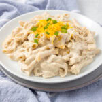 A white plate filled with creamy pasta garnished with shredded cheddar cheese and chopped green onions. The pasta, covered in a thick white sauce, is served on a light gray tablecloth.