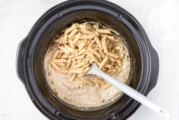 A slow cooker filled with a creamy sauce and uncooked penne pasta, with a white spoon resting in the mixture, ready for cooking.