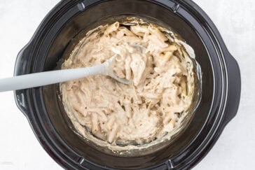 A crockpot filled with creamy, cooked pasta. A gray spoon is resting in the pasta, which is coated in a thick, white sauce. The crockpot is placed on a white surface.