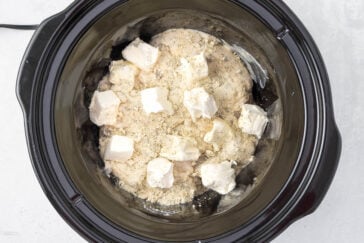 A slow cooker filled with partially cooked dough, topped with cubes of cream cheese and sprinkled with breadcrumbs, ready for cooking. The surface beneath is light gray.