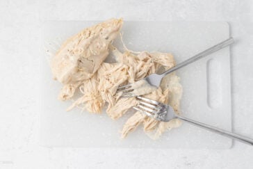Two forks shredding cooked chicken breast on a white cutting board. The chicken is covered in a creamy sauce.
