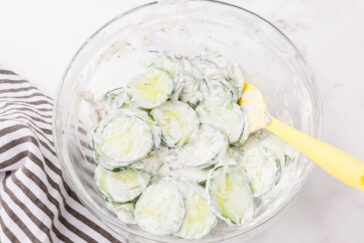 A glass bowl filled with creamy cucumber salad, featuring sliced cucumbers coated in a white dressing. A yellow spoon rests inside the bowl. A striped black and white cloth is partially visible on the left.