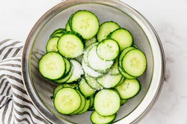 A metal colander containing sliced cucumbers sprinkled with salt, placed on a marble countertop. A striped cloth is partially visible on the side.