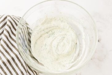 A glass bowl filled with creamy ranch dressing, lightly speckled with herbs. A striped cloth napkin is partially visible to the left. The background is a light marble surface.